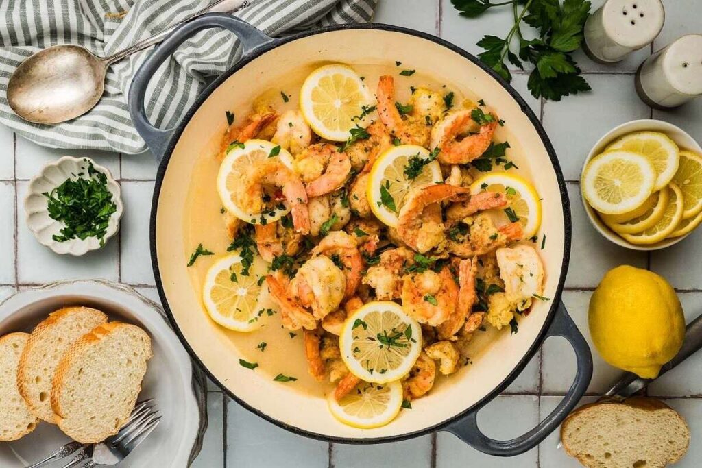 A skillet of Shrimp Francese with lemon slices and parsley, surrounded by bread, lemon, and fresh herbs on a table.