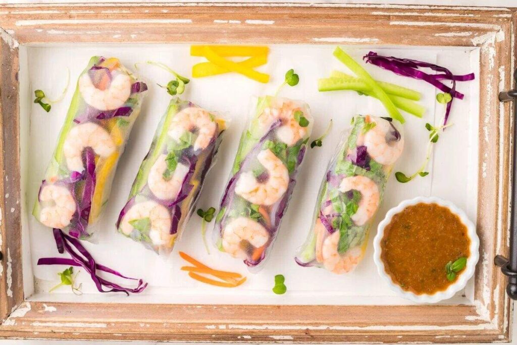 Four shrimp spring rolls with dipping sauce and sliced vegetables on a white tray.