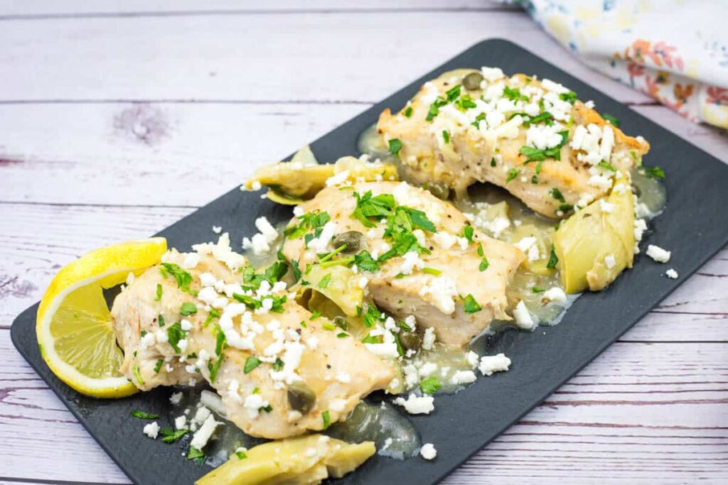 Three pieces of baked chicken topped with herbs and crumbled cheese are served with artichoke hearts and lemon slices on a black rectangular plate.