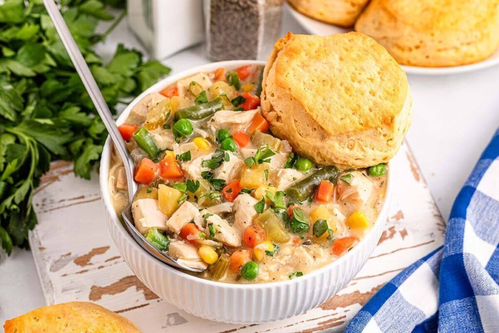  A bowl of slow cooker chicken pot pie soup with vegetables and a biscuit on top rests on a rustic wooden surface.