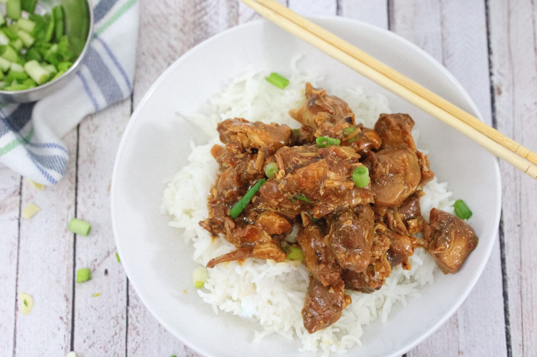 White bowl with rice and teriyaki chicken topped with green onions with chop sticks on the bowl and more green onions behind it.
