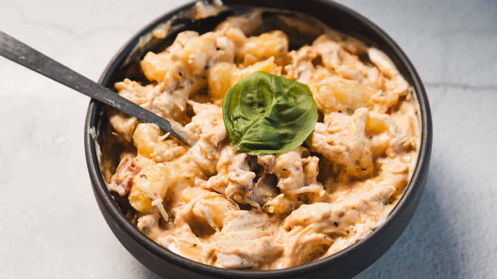 A black bowl filled with creamy pasta coated in a thick sauce, topped with a fresh basil leaf. A metal spoon rests inside the bowl. The background is a light-colored surface.