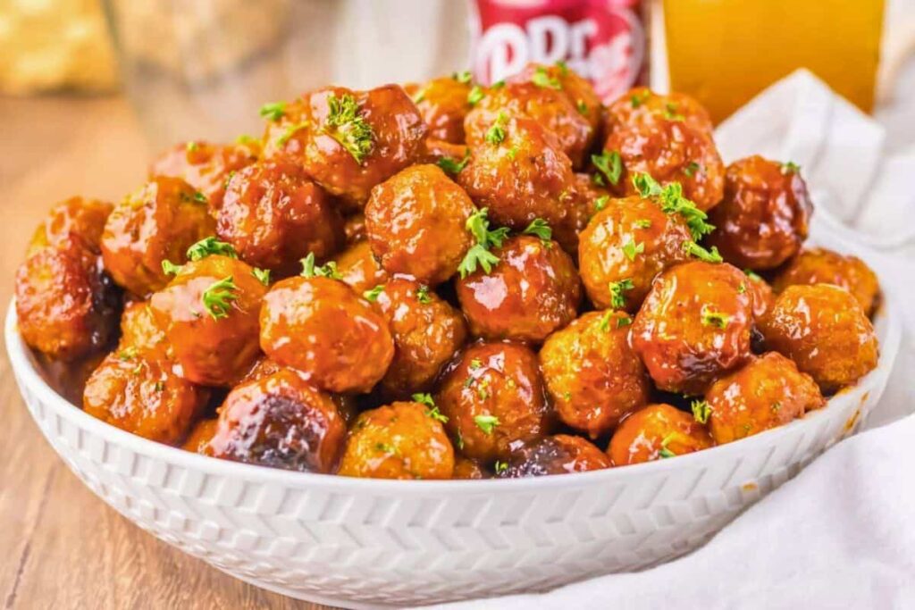 A white bowl filled with glazed meatballs, garnished with chopped parsley, placed on a wooden table with a soft drink can and a glass in the background.