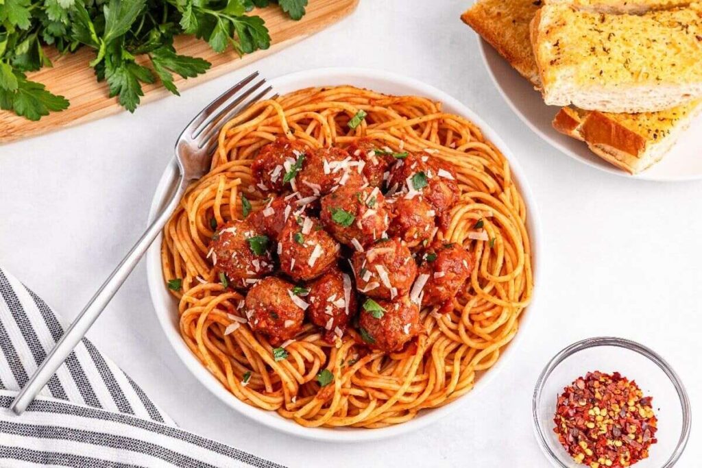Spaghetti with meatballs, cheese, and herbs, served with garlic bread and crushed red pepper.