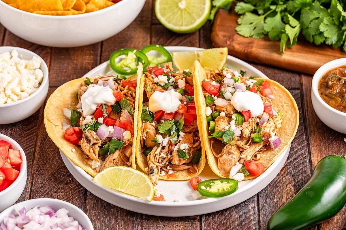 Three chicken tacos with toppings including sour cream, diced tomatoes, onions, jalapeños, and cilantro on a white plate, garnished with lime wedges. 