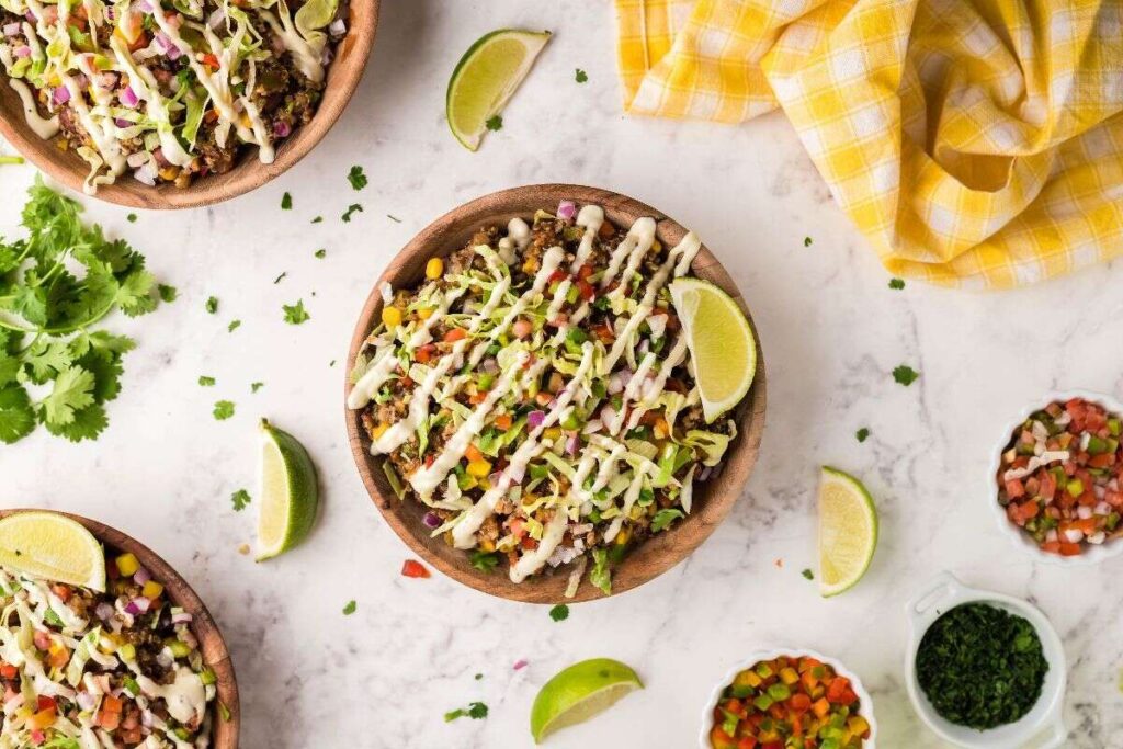 A southwest taco bowl with creamy dressing, lime wedges, and colorful veggies sits on a marble surface, perfect alongside Crockpot Mexican quinoa.