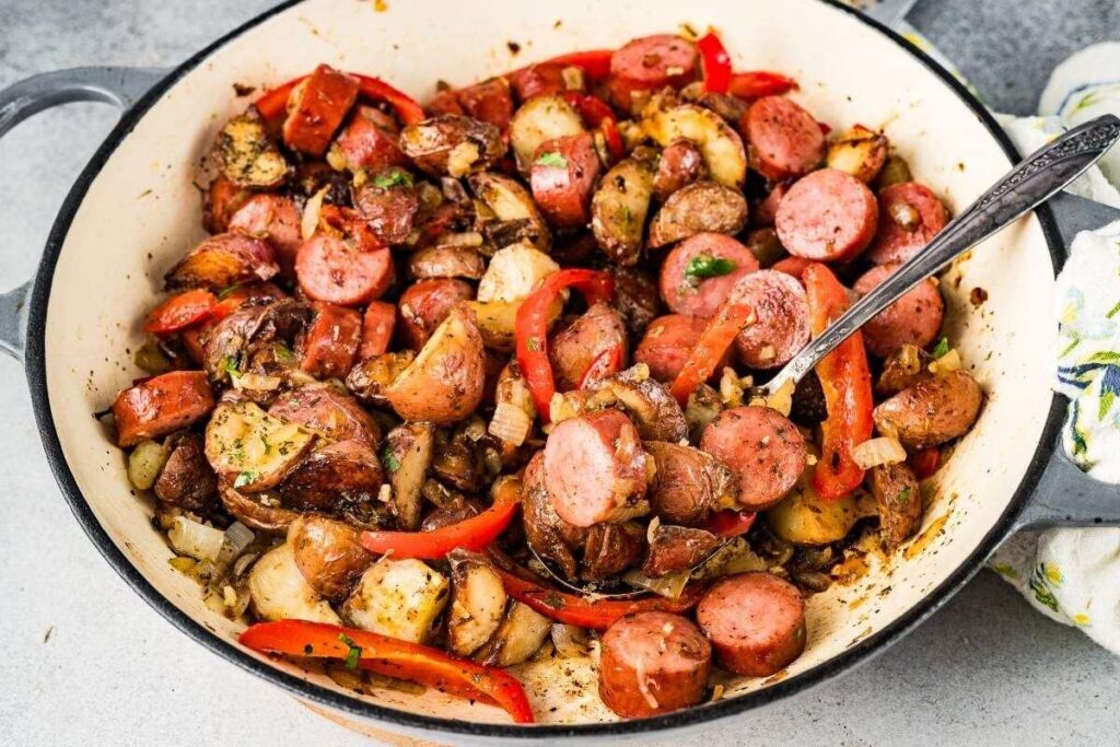A smoked sausage and potato skillet filled with sautéed sliced sausage, red bell peppers, onions, and diced potatoes, stirred together with a metal spoon.