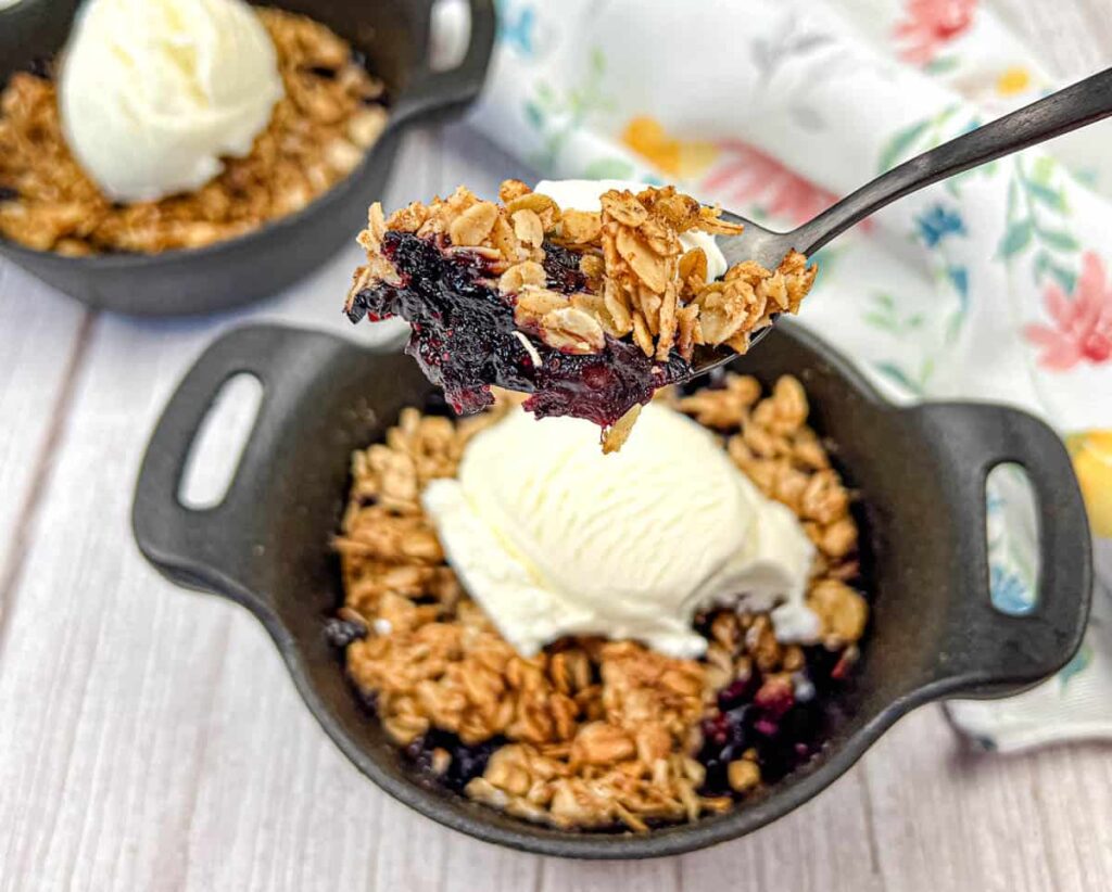 Close-up shot of individual blueberry crumble desserts in black cast iron skillets, each topped with a scoop of vanilla ice cream, with a spoonful of the dessert being lifted up, displaying the layered texture of the crumble and filling.