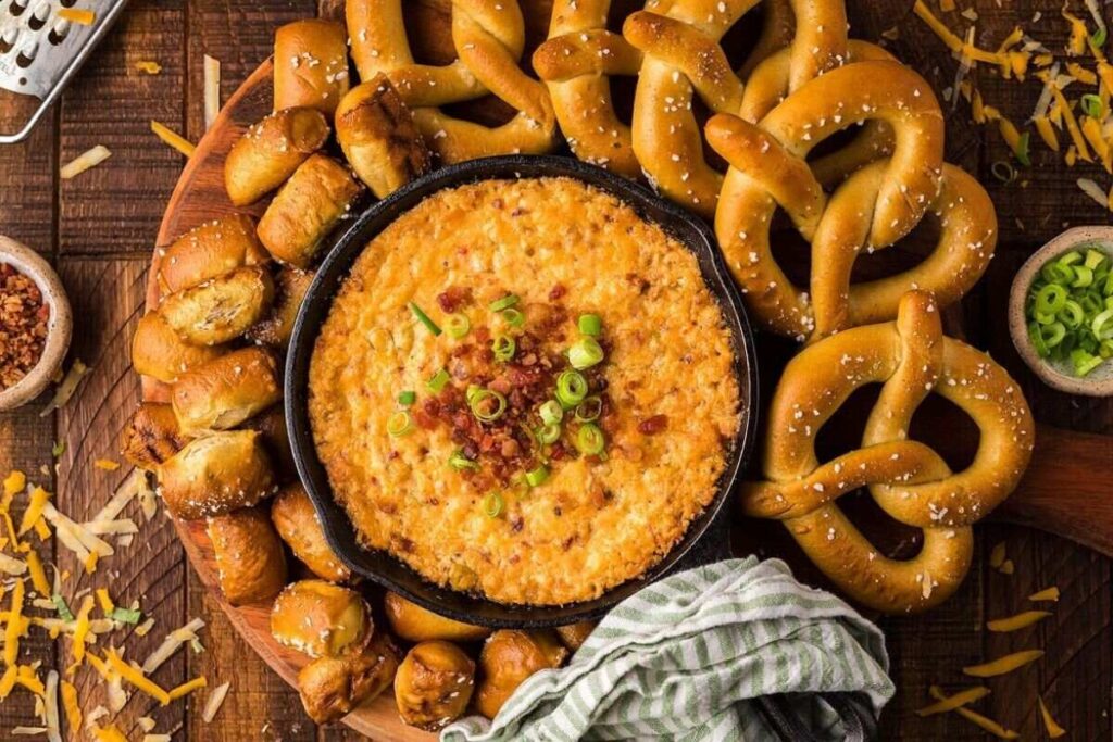 Smoked gouda and cheddar bacon dip topped with green onions and crispy bacon, served with pretzels on a wooden board.