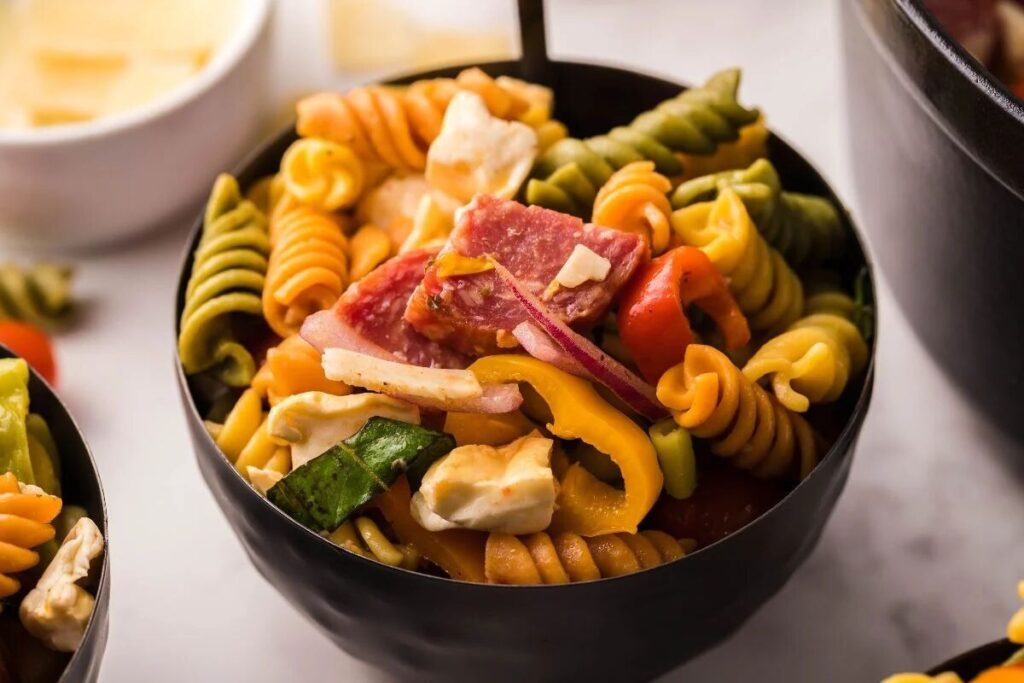 A black bowl of colorful pasta salad with cheese, salami, veggies, and zesty Pepperoncini dressing, perfect for summer BBQs.