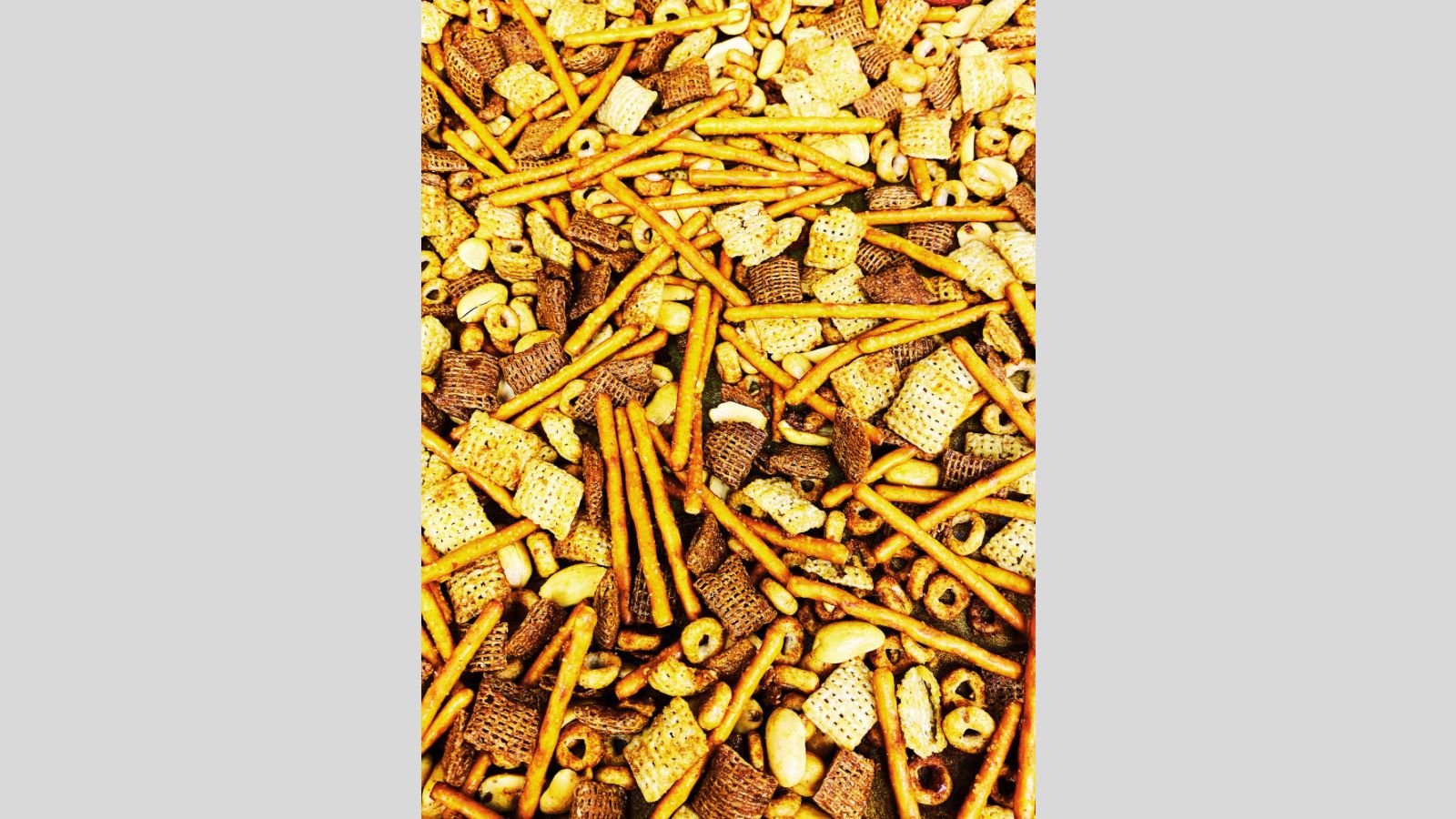 A close-up view of a crunchy snack mix featuring pretzel sticks, cereal squares, oat rings, peanuts, and other bite-sized pieces. The well-seasoned snack mix looks perfect for on-the-go munching.