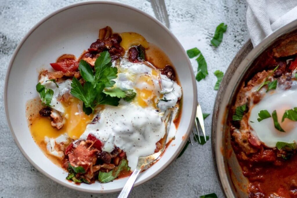 A bowl of shakshuka with spinach and red beans, topped with a runny egg, yogurt, and fresh parsley.