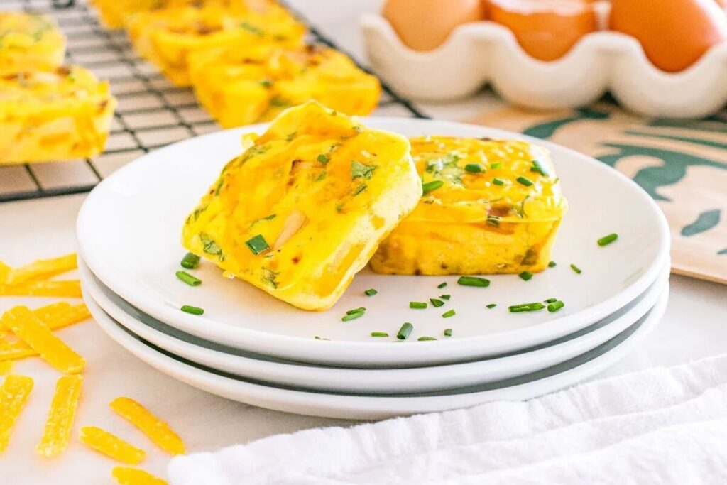 Two baked egg bites with herbs sit on a white plate, with eggs and muffins blurred in the background.