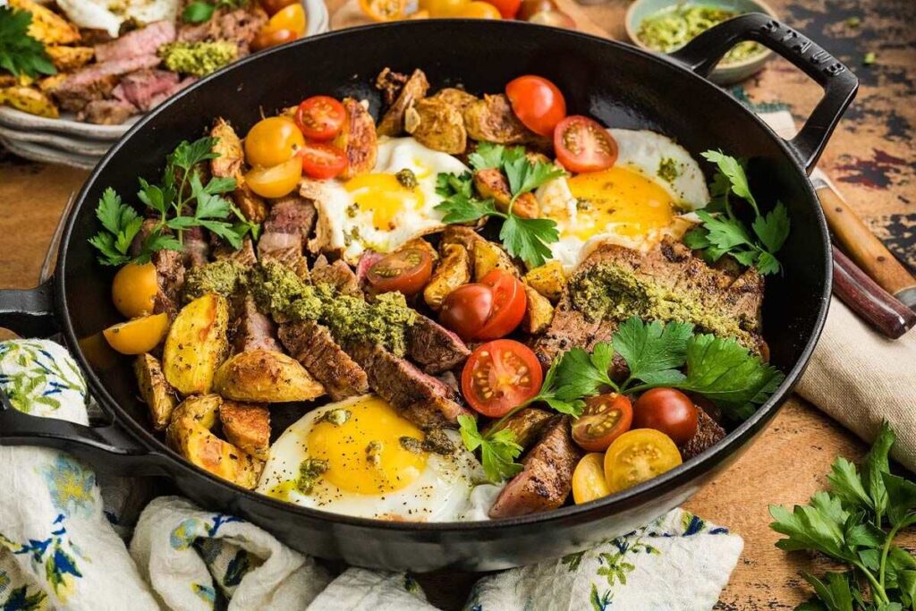 Hearty skillet with steak, eggs, roasted potatoes, tomatoes, fresh herbs, and green sauce.