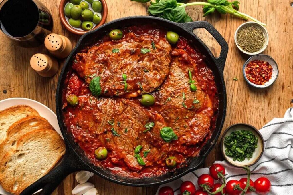 Steak in tomato sauce with olives and basil, served with bread and herbs.