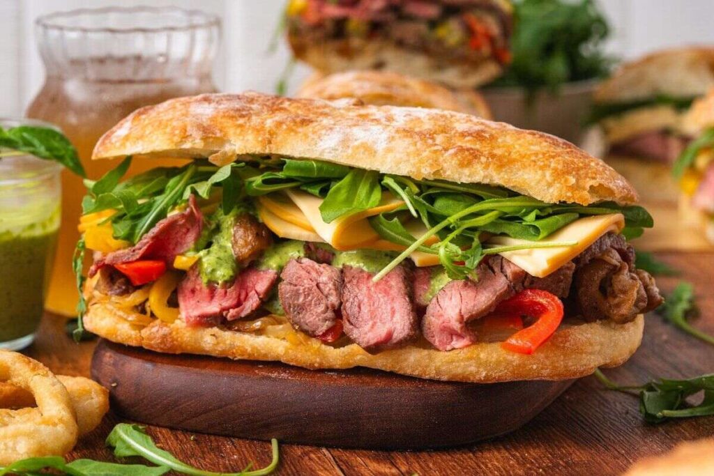 Steak sandwich with cheese, arugula, red peppers, and sauce on ciabatta, served on a wooden board.