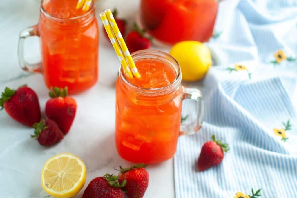 Mason jars filled with iced strawberry lemonade, garnished with yellow striped straws, surrounded by strawberries and a halved lemon on a light surface.
