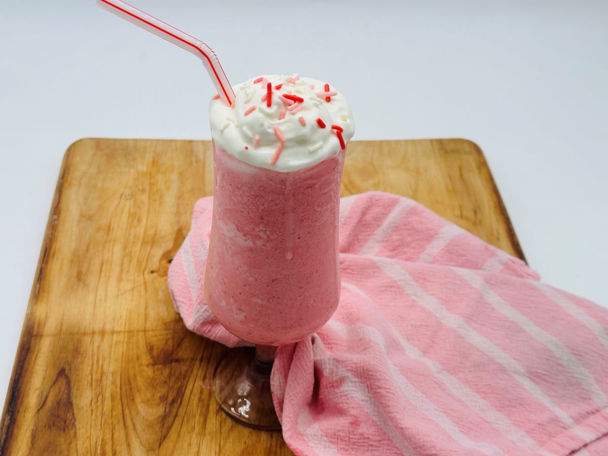 A pink milkshake topped with whipped cream and sprinkles, served with a red-striped straw on a wooden board and a pink striped cloth.