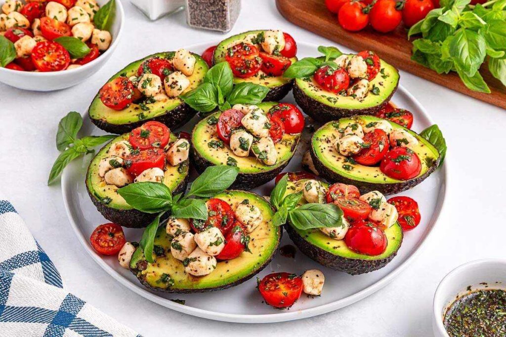 Halved avocados filled with cherry tomatoes, mozzarella, and basil, garnished with herbs, on a white plate.