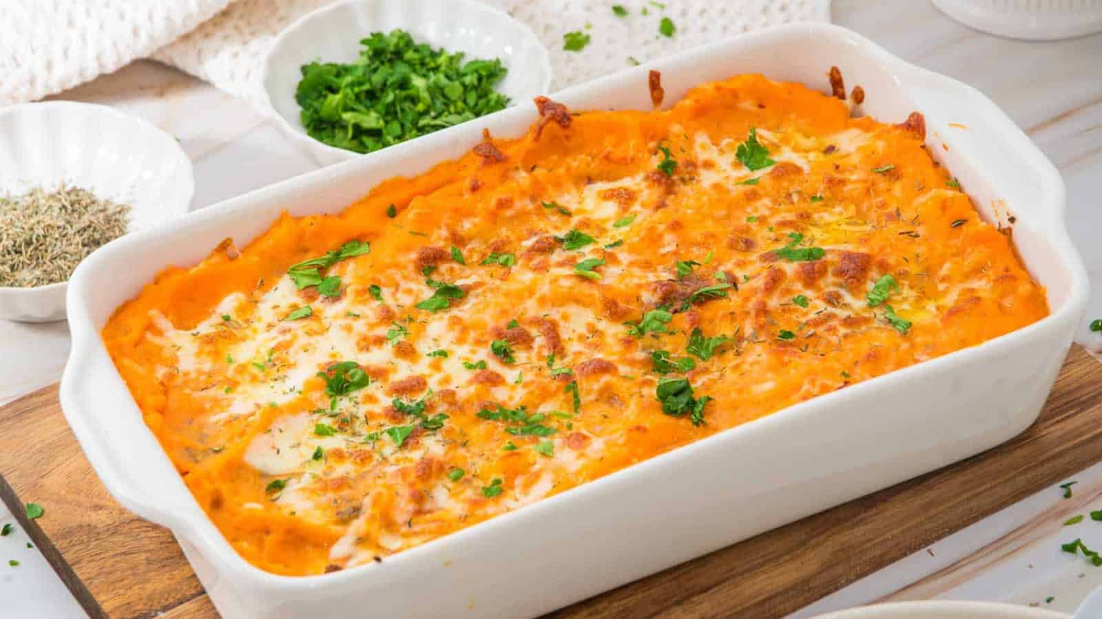 A baked sweet potato casserole topped with melted cheese and chopped parsley, served in a white rectangular dish on a wooden board. Small bowls of fresh herbs and seasonings are visible in the background.