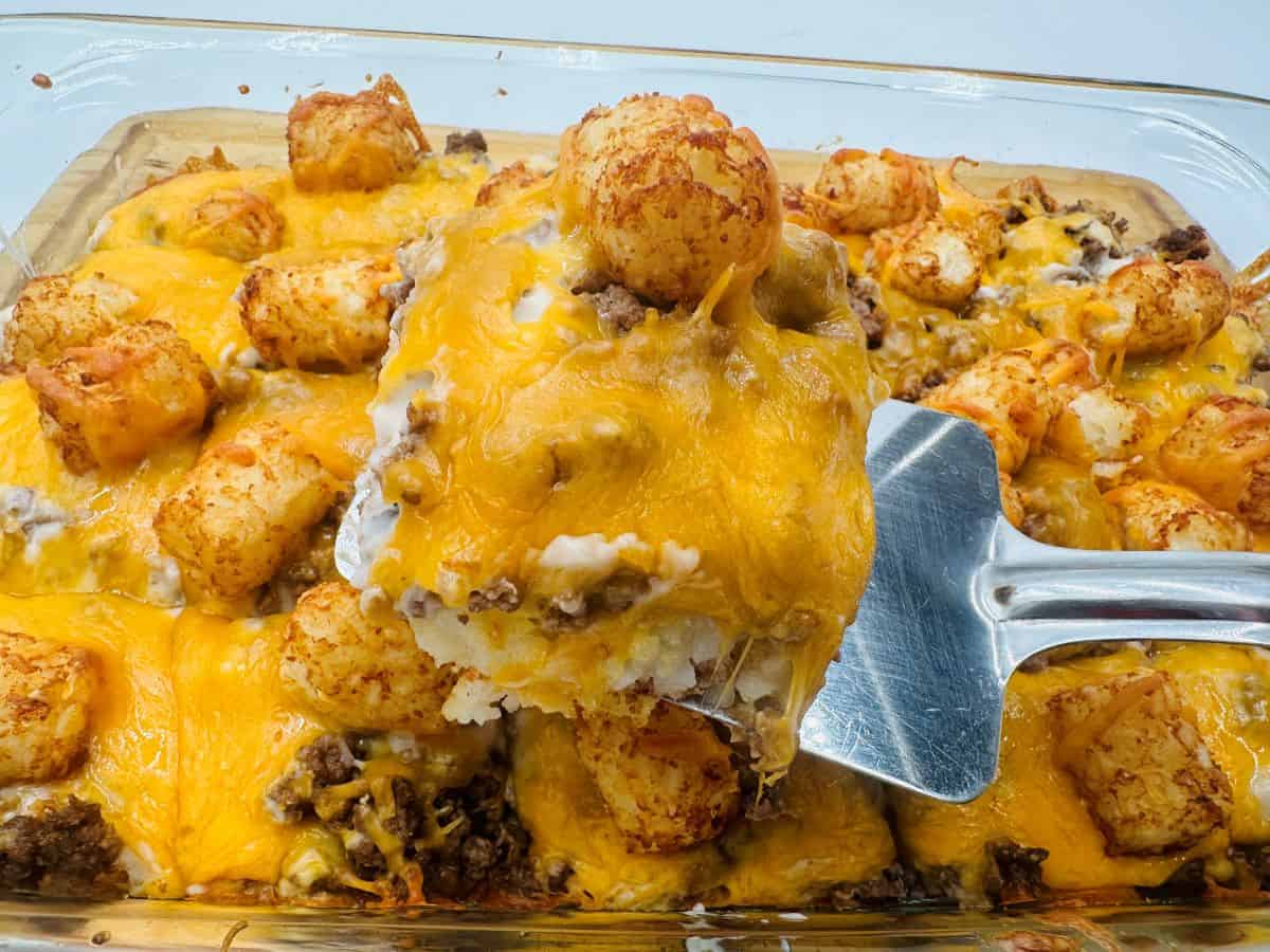 A spatula lifts a portion of a baked casserole featuring tater tots, ground beef, and melted cheddar cheese from a glass dish.