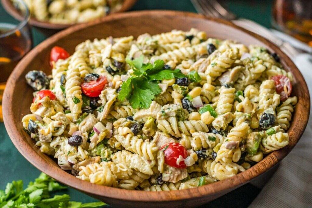 A wooden bowl filled with southwest pasta salad, rotini, tomatoes, olives, and corn, topped with tender Tex Mex chicken and fresh herbs.