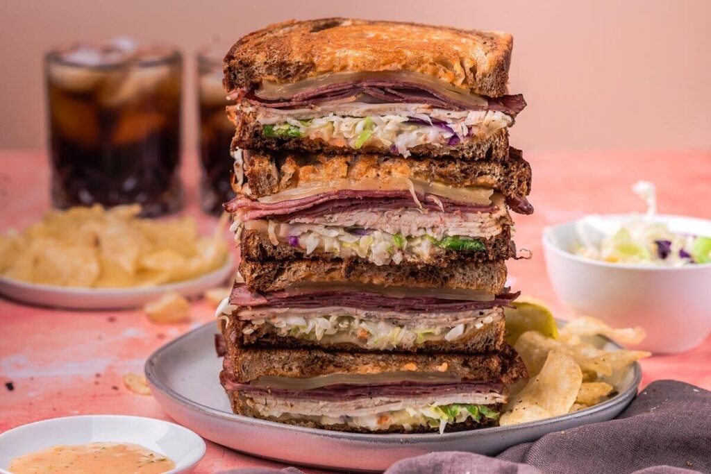 A stacked Rachel sandwich with layers of meat, cheese, lettuce, and sauce on toasted bread is served on a plate with potato chips. 