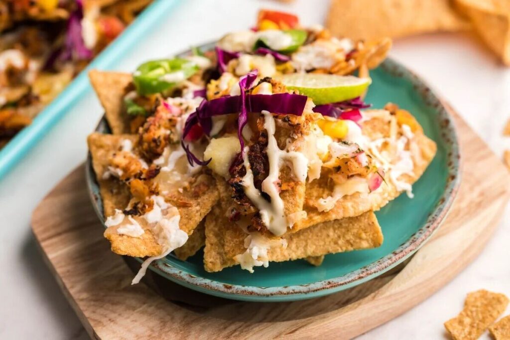 Grilled tilapia nachos with tortilla chips, shredded cabbage, zesty sauce, and lime slices on a teal plate.