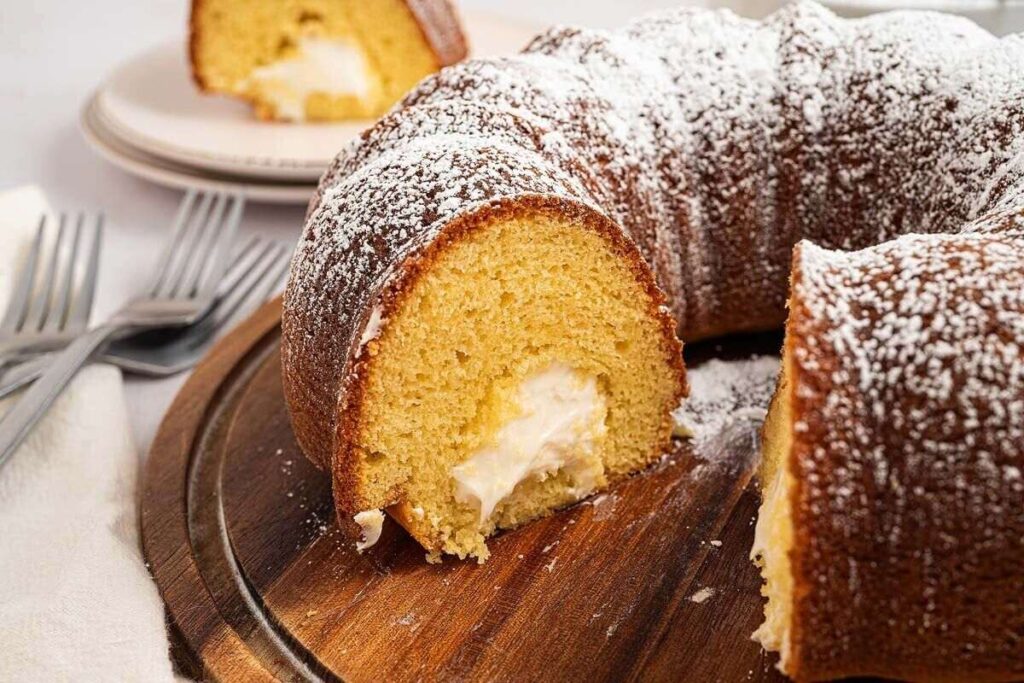 A sliced Twinkie Cake Bundt with cream filling and powdered sugar on top, served on a wooden board.
