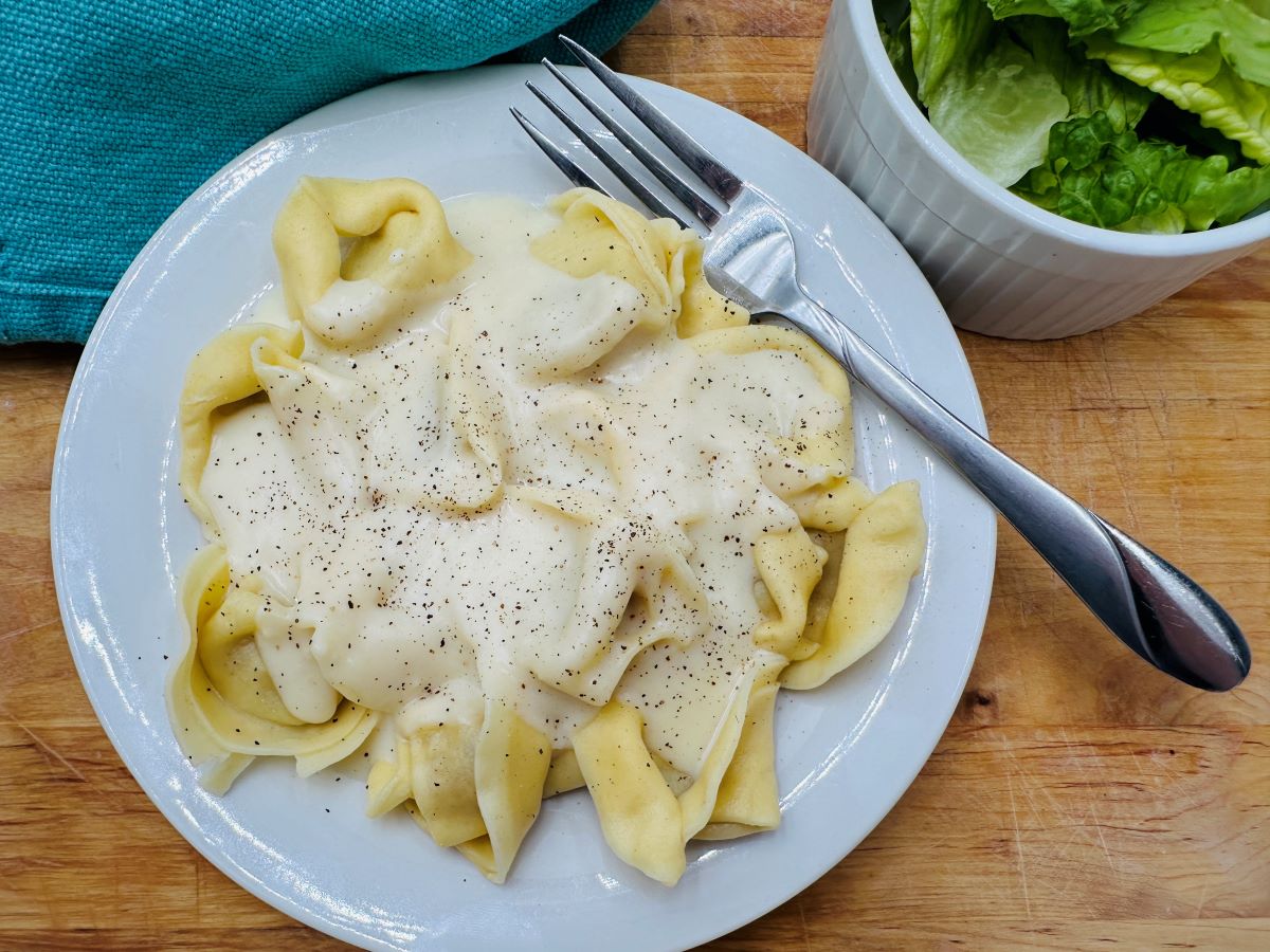 A plate of tortellini covered in a creamy white sauce, garnished with black pepper, next to a fork and a small bowl.