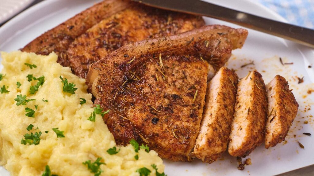A plate of sliced seasoned pork chops served with creamy mashed potatoes, garnished with parsley.
