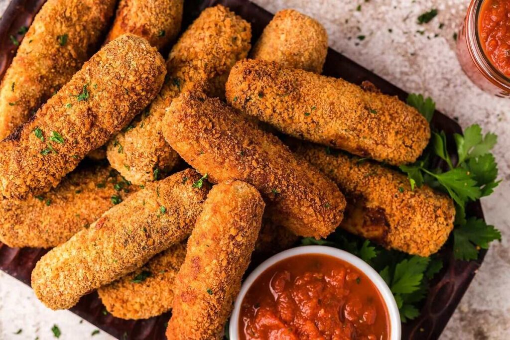 Crispy mozzarella stick chicken fries on a tray with parsley, served with a bowl of red dipping sauce.