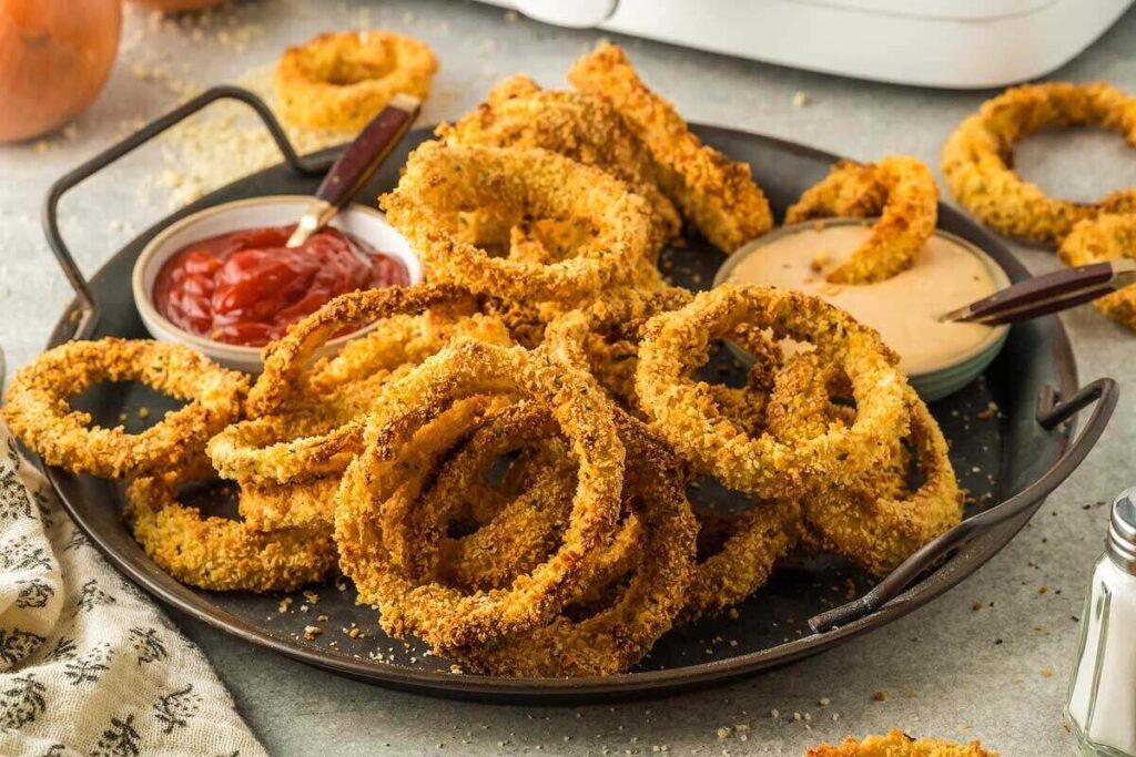 A plate of crispy air fryer onion rings served with ketchup and dipping sauce on a gray tablecloth.