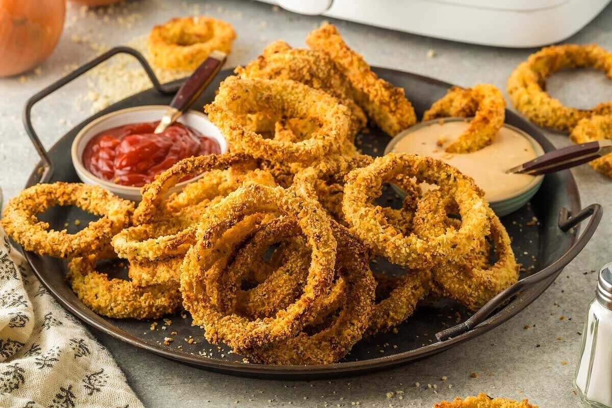 A serving of crispy fried onion rings on a dark plate, garnished with chopped herbs, with dipping sauce and chopped green onions in small glass bowls.