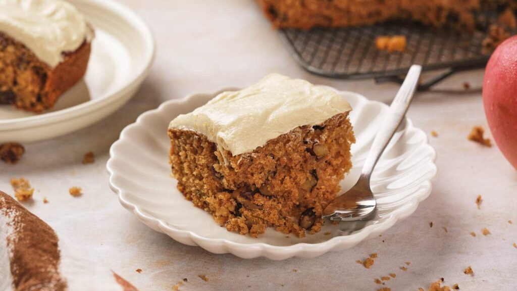 A slice of cake with frosting on a white plate, with a fork beside it.