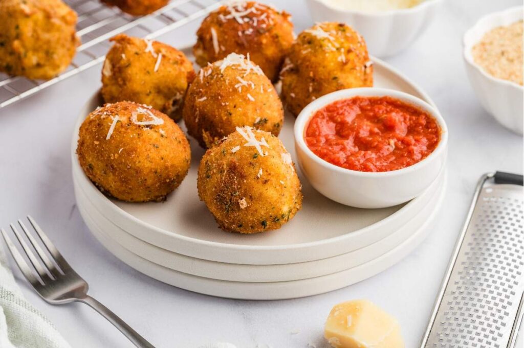 Arancini with grated cheese served alongside marinara sauce, with a fork and cheese nearby.