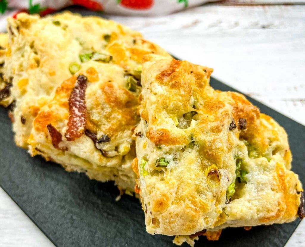 Close-up shot of two golden-brown baked savory biscuits, studded with green onion and bacon bits, resting on a black slate surface.