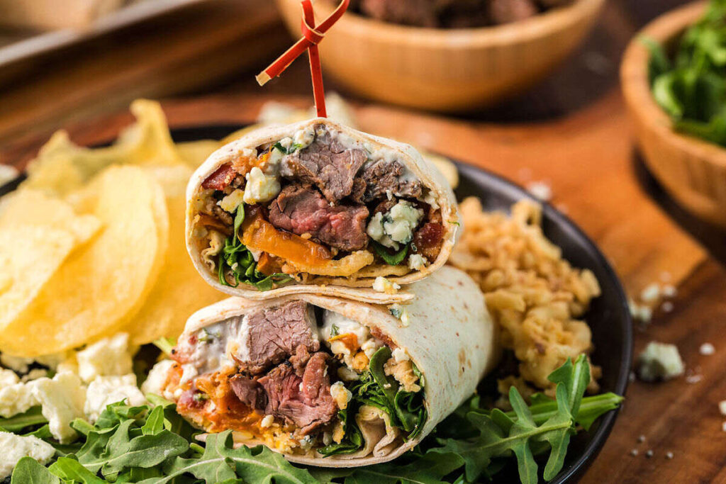 A close-up of a sliced wrap filled with beef, blue cheese, lettuce, and other ingredients, garnished with chips and greens on a plate.
