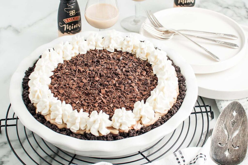 No Bake Baileys Cream Pie topped with whipped cream and chocolate shavings, with plates and utensils in the background.