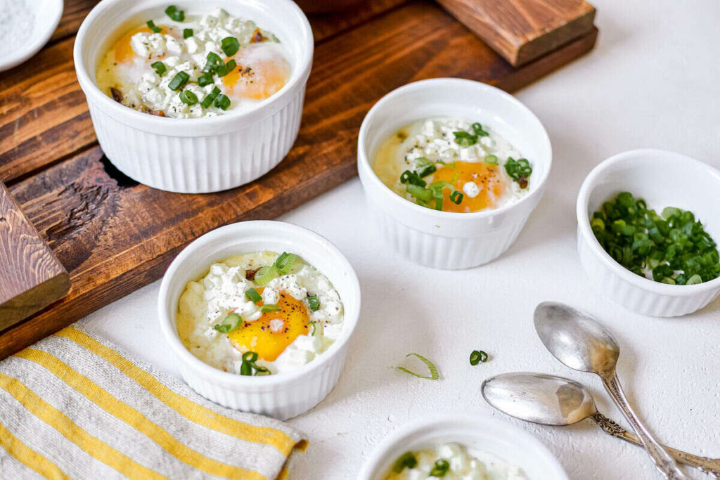 Baked eggs with cheese and green onions served in white ramekins, with spoons and a bowl of chopped green onions nearby.