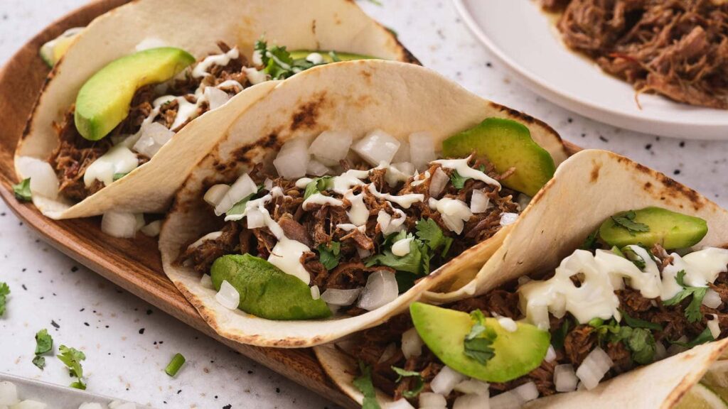 Barbacoa tacos on a long wooden plate.