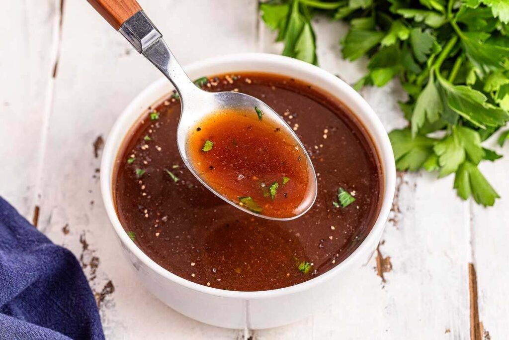 Spoon above a bowl of rich brown beef au jus with herbs.