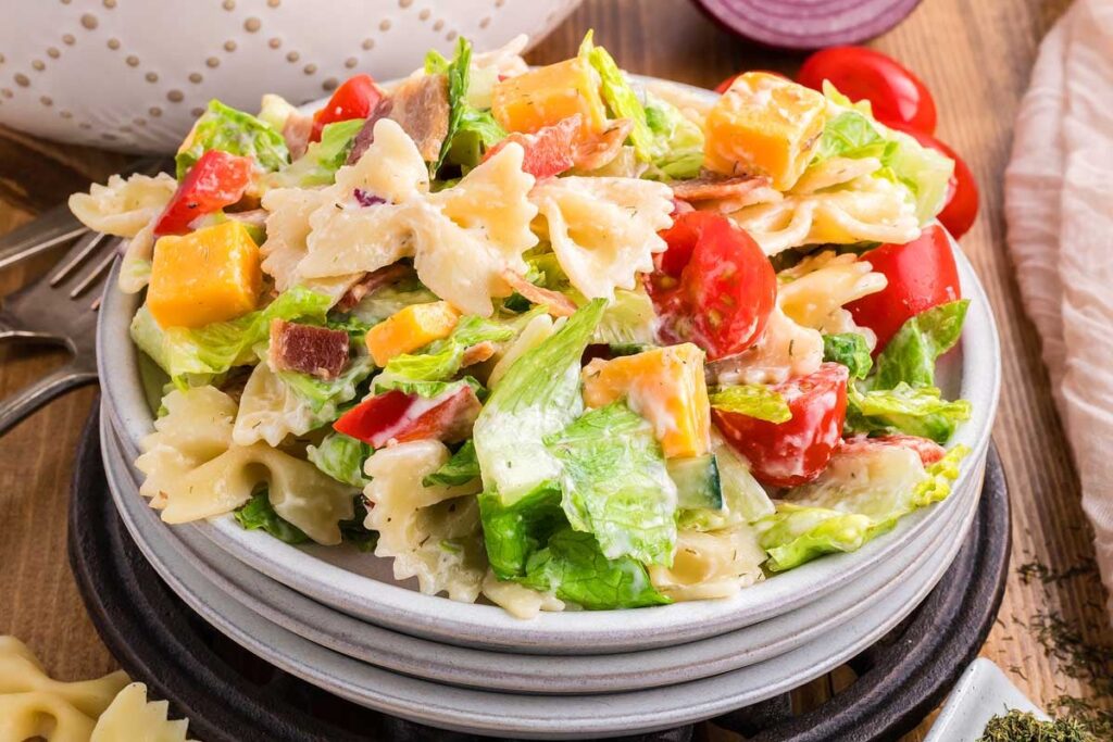 A bowl of BLT pasta salad with bowtie pasta, lettuce, tomatoes, cheese cubes, bacon, and dressing on a wooden table.