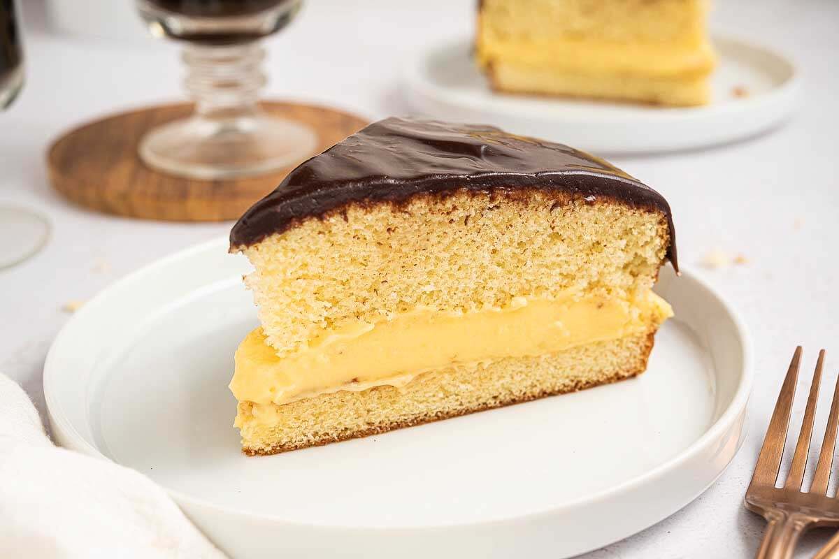 A close-up of sliced Boston cream pie showing yellow sponge cake layers filled with creamy custard and topped with glossy chocolate glaze.