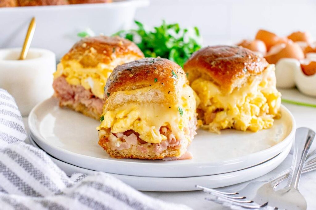 Three Hawaiian roll breakfast sliders with scrambled eggs, ham, and cheese sit on a plate, garnished with parsley.