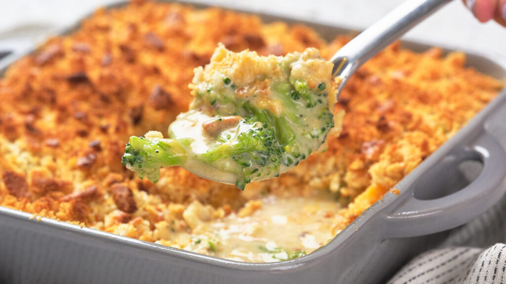A person's hand lifting a portion of broccoli casserole with a spoon.