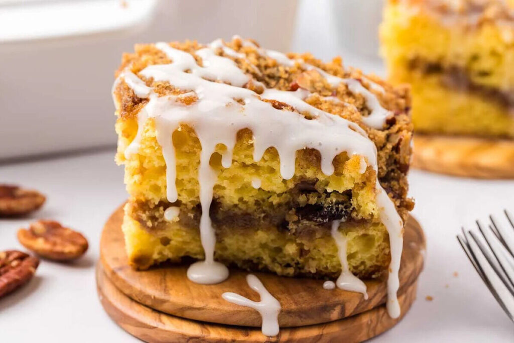 A slice of cake mix coffee cake with a pecan crumble and white glaze on a wooden plate, with more cake and pecans in the background.