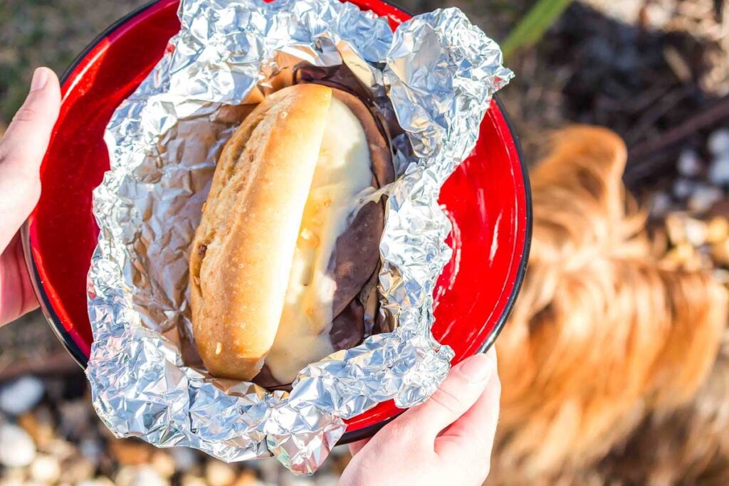 A red plate with a campfire foil pack French dip sandwich on it at the campfire.