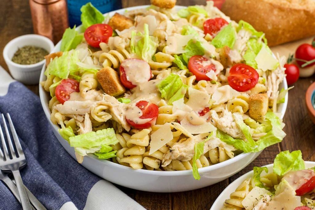 A bowl of chicken Caesar pasta salad with rotini, cherry tomatoes, lettuce, shredded chicken, croutons, and parmesan, served with a fork and napkin.