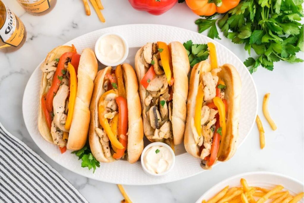 Four chicken Philly sandwiches garnished with bell peppers and parsley, served with dips on an oval white plate, surrounded by fries, a striped napkin, and fresh vegetables.